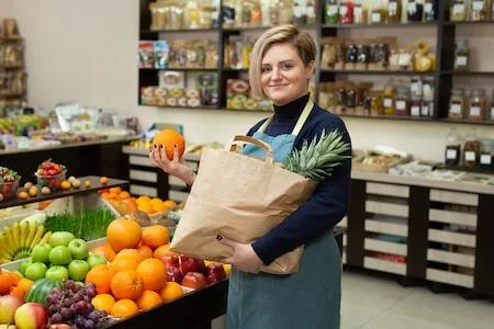 Eine Person in einer Schürze steht in einem Lebensmittelgeschäft und hält eine Papiertüte mit einer Ananas in der Hand, während sie in der anderen Hand eine Orange hält. Auf der Theke sind verschiedene Früchte ausgestellt.