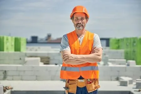 Ein Bauarbeiter in einer orangefarbenen Sicherheitsweste und einem Schutzhelm steht mit verschränkten Armen auf einer Baustelle.