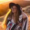 Eine Frau mit langen Haaren, einem breitkrempigen Hut und einer Brille steht auf einem sonnenbeschienenen Feld und hält eine bunte Decke in der Hand.